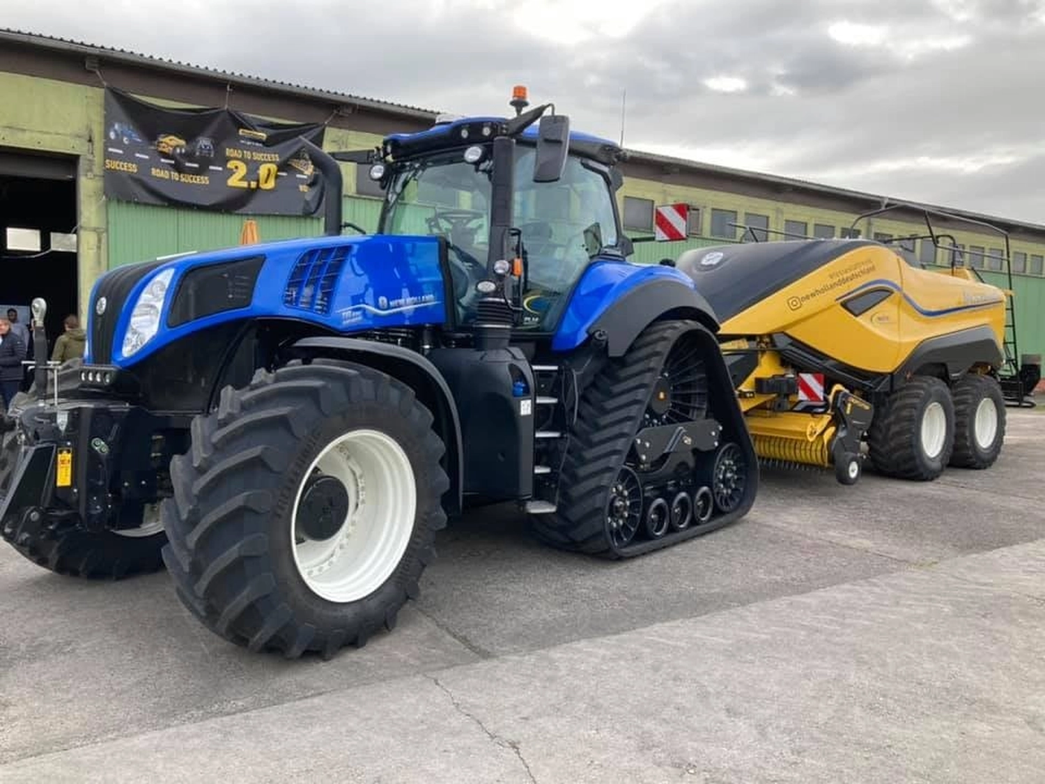 NM- Nerchau-Mutzschener - Landwirtschaft - Ein blauer New Holland T8 SmartTrax-Traktor mit Vorderreifen und Raupenketten zieht eine gelbe Ballenpresse auf einem gepflasterten Parkplatz.